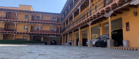 Un patio con piso de piedra, un edificio con varios balcones y una escalera, y varios coches aparcados.