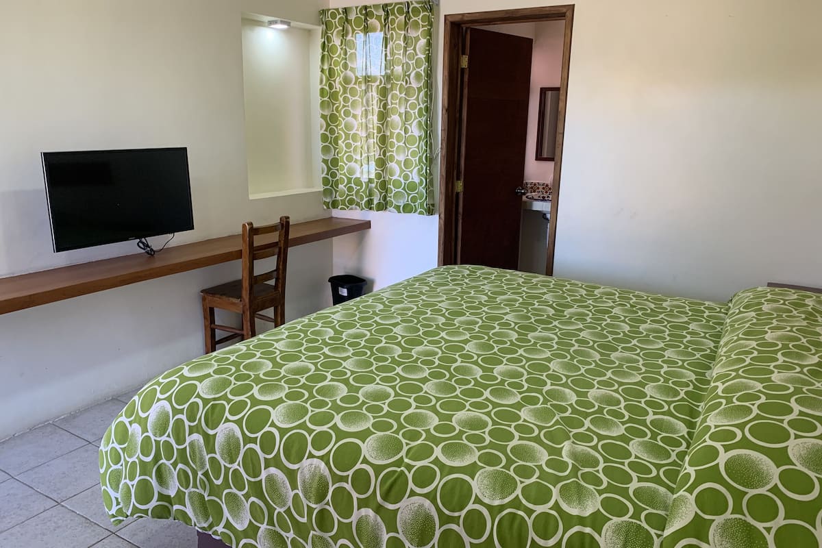 A bedroom with a bed, a TV on a shelf, a chair, a door, and a bathroom visible through an open door.