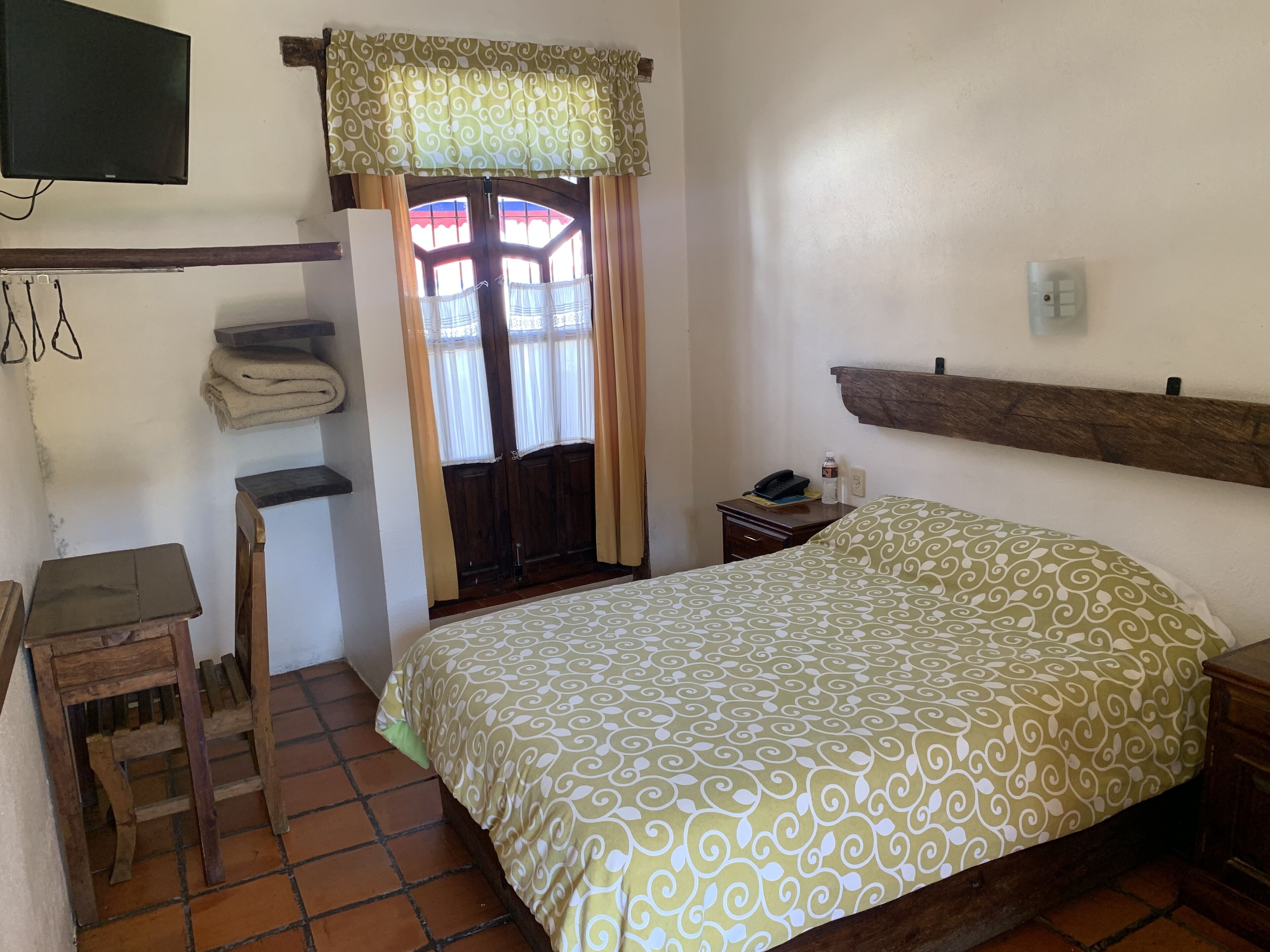 Un dormitorio con una cama, una mesita de noche, una silla de madera, un televisor fijado en la pared y una ventana con cortinas.