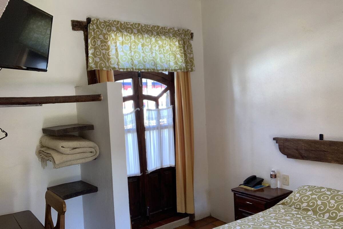A room with a bed, a desk, a chair, a shelf with towels, a small bedside table with a phone and bottle, and a window with curtains.
