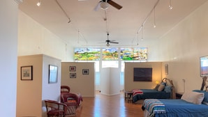 A spacious room with a large bed, two smaller beds, a ceiling fan, and a large window with a colorful view. - Former Art Gallery is now spacious lodging (Sedan)