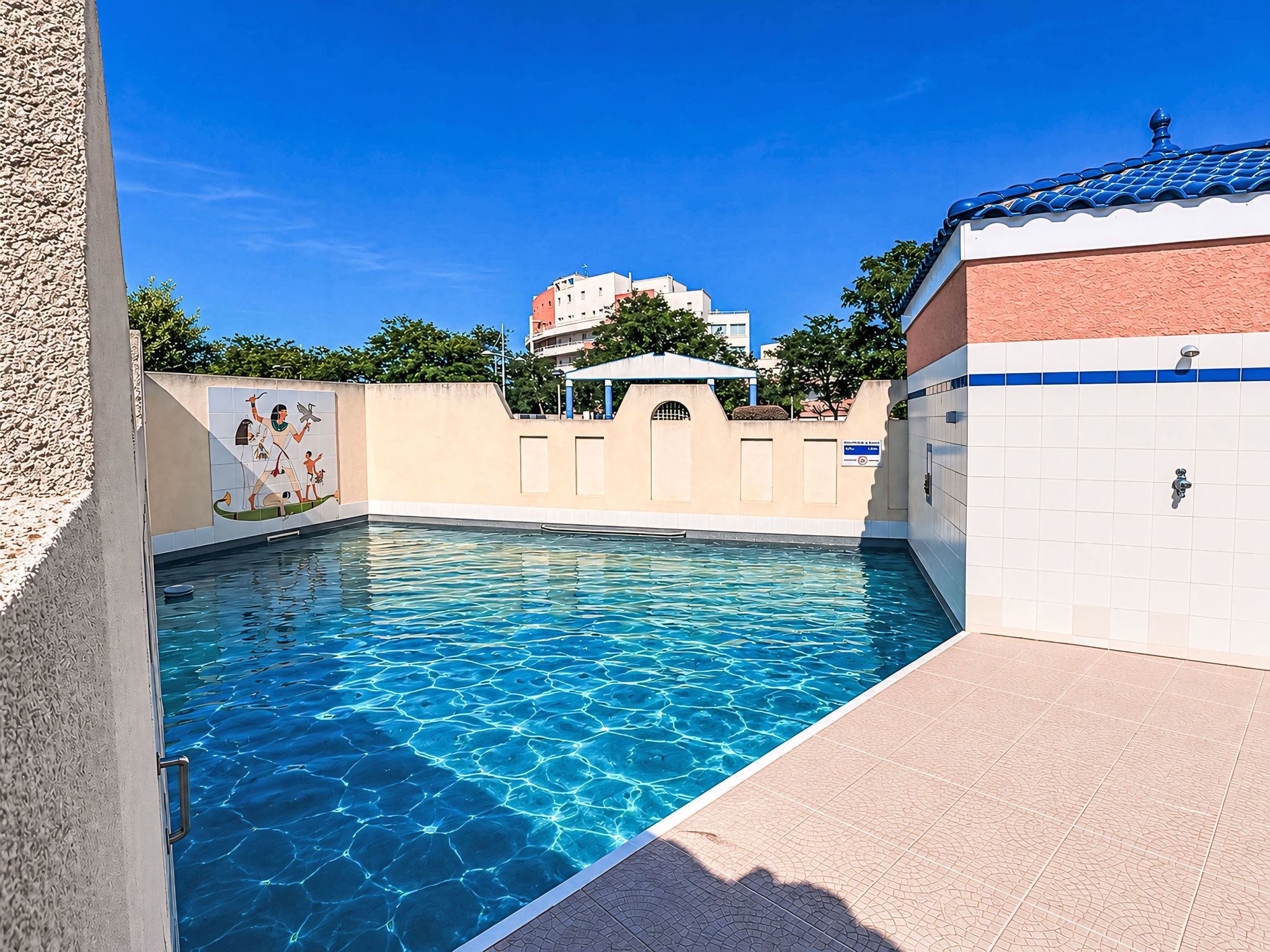 A swimming pool with clear blue water, a tiled wall with a mural, and a building with a blue roof in the background.