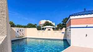 Uma piscina com água azul cristalina, uma parede revestida com um mural, e um edifício com telhado azul ao fundo.