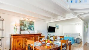 Uma área de refeições com mesa e cadeiras de madeira, uma fruteira e uma televisão ao fundo.