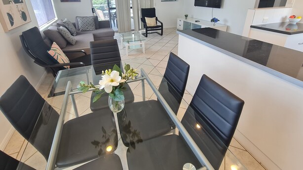 A modern living room with a glass dining table, black chairs, a sofa with patterned cushions, a TV mounted on the wall, and a kitchen area with a black countertop and white cabinets. - Cranbourne Court Beach Side Apartments (Maroochydore)
