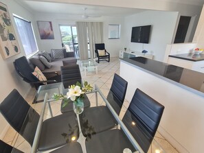 A modern living room with a glass dining table, black chairs, a sofa with patterned cushions, a TV mounted on the wall, and a kitchen area with a black countertop and white cabinets. - Cranbourne Court Beach Side Apartments (Maroochydore)