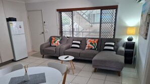 A modern living room with a grey sofa, a dining table with chairs, a white refrigerator, and a window with blinds. - Cranbourne Court Beach Side Apartments (Maroochydore)