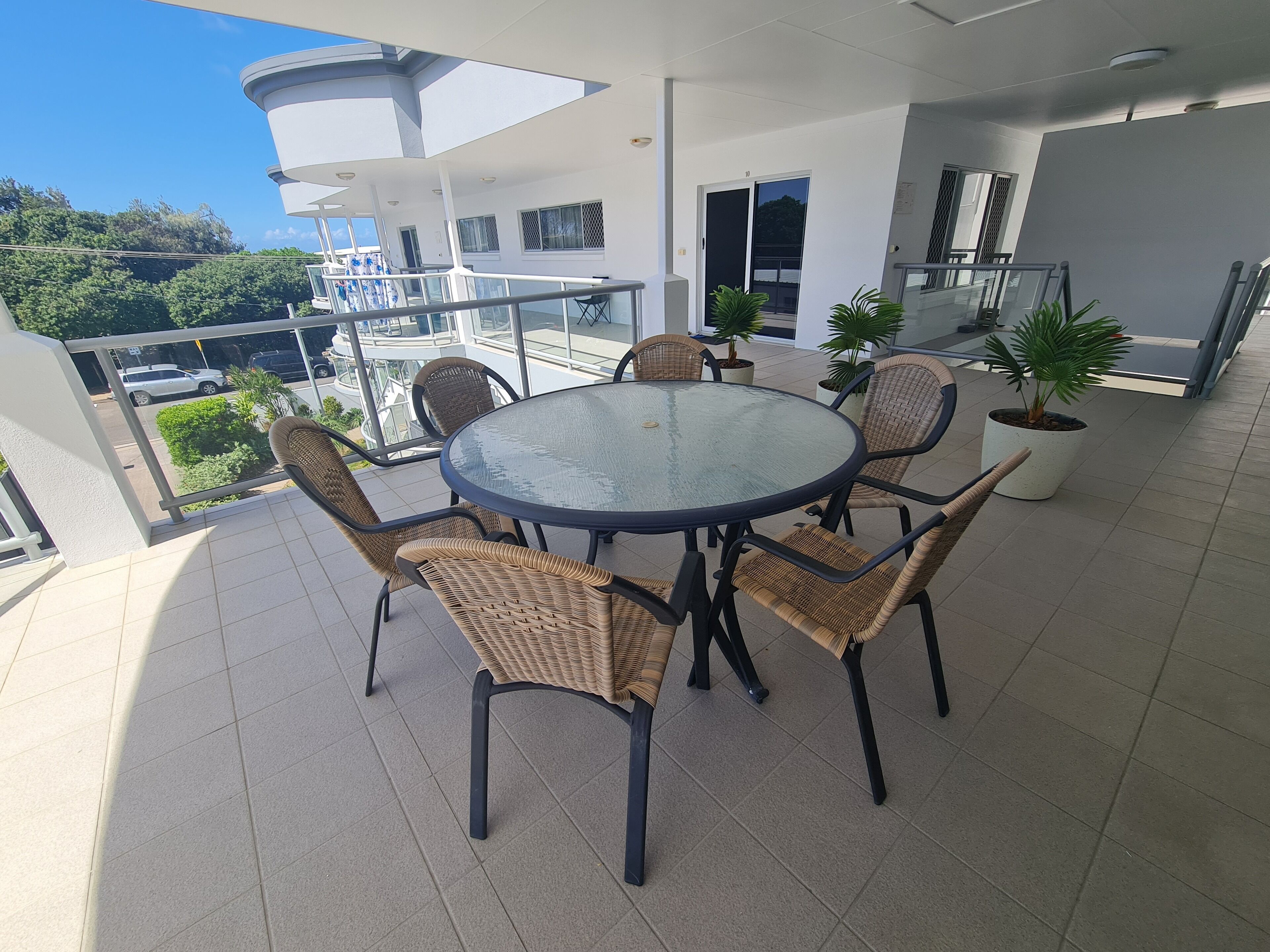 A balcony with a round glass table and wicker chairs, surrounded by potted plants.