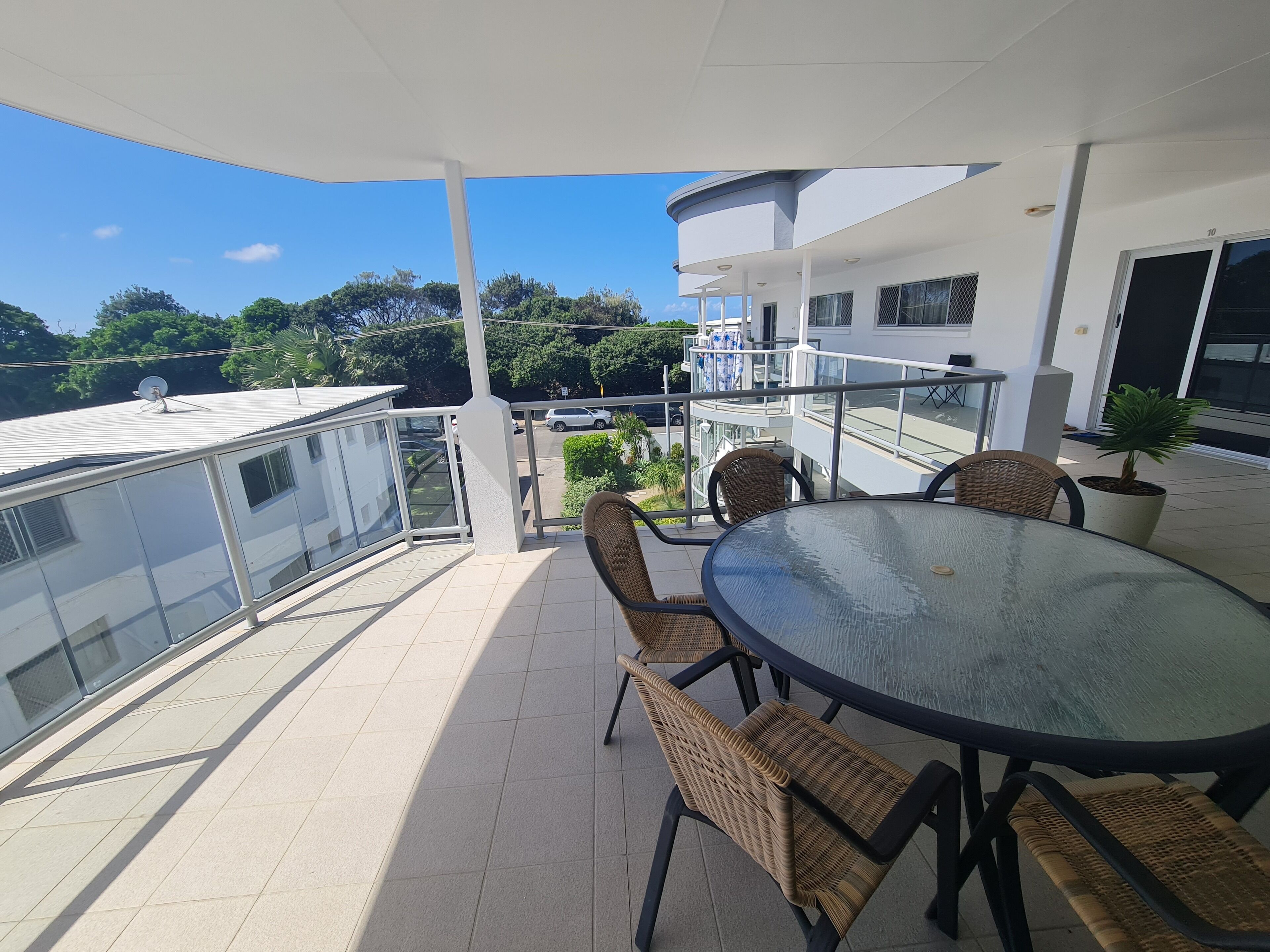 A balcony with a glass table and wicker chairs, overlooking a parking area and greenery.