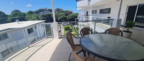 A balcony with a glass table and wicker chairs, overlooking a parking area and greenery.
