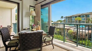 A balcony with wicker furniture, a table with a wine glass, and a view of a hotel building and pool. - Waipouli Beach Resort B202 - Resort Pool, Waterslides, AC, & Steps to Beach Fun! (Kapaa)
