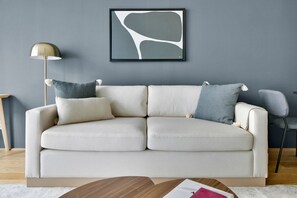 Ett modernt vardagsrum med en beige soffa, två kuddar, en inramad abstrakt målning, en golvlampa och ett soffbord i trä.