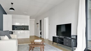 Un salon moderne avec un téléviseur à écran plat, une table basse et un canapé. Au fond, on aperçoit un coin cuisine avec des meubles blancs et une table à manger.