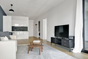 A modern living room with a flat-screen TV, a coffee table, and a sofa. There is a kitchen area in the background with white cabinets and a dining table.