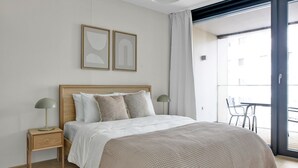 Une chambre à coucher comprenant un lit, deux tables de chevet, une commode, un luminaire au plafond et donnant sur le balcon extérieur.