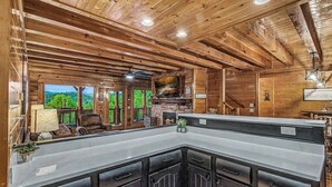 L’intérieur d’un chalet en bois, avec une cheminée en pierre, un grand comptoir de cuisine et une vue sur l’extérieur.
