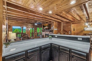 Ein Holzblockhaus-Interieur mit Kamin aus Stein, einer großen Küchenzeile und Blick ins Freie.