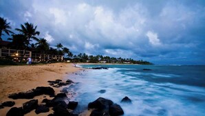 A beach with a rocky shoreline, palm trees, and a building with lights in the background. - 10 Bedroom Sleeps 24 w/ AC - Less than a 1 Minute Walk with views of Poipu Beach (Koloa)