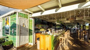 Un área de bar al aire libre con una decoración verde y blanca, mobiliario de madera y luces colgantes.