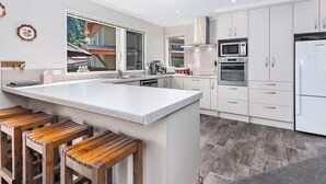 A modern kitchen with white cabinets, a large island with stools, a microwave, oven, and refrigerator. - Denbigh Escape - Perfect location, family-friendly! (Arrowtown)