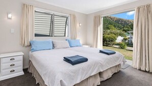 A bedroom with a large bed, two bedside tables, a window with curtains, and a view of a green landscape. - Denbigh Escape - Perfect location, family-friendly! (Arrowtown)