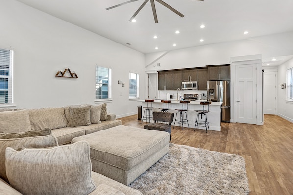 A spacious living room with a large sectional sofa, a small ottoman, and a fluffy rug. There is a kitchen area with modern appliances and bar stools.