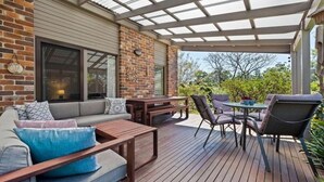 Um pátio externo com deck de madeira, área de estar com almofadas e uma mesa com cadeiras.
