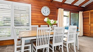 Uma sala de jantar com uma mesa de madeira e cadeiras brancas, um relógio na parede e uma janela com persianas.