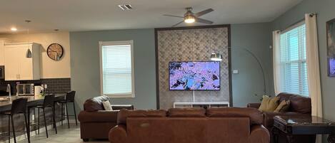 Una sala de estar moderna con un sofá marrón, un televisor montado en la pared y una zona de comedor con una cocina al fondo.