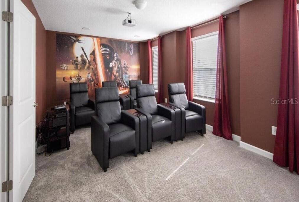 Una sala de cine en casa con cuatro sillones reclinables de cuero negro, un cartel de película grande en la pared y piso alfombrado.