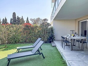 Une terrasse avec une table et des chaises, un petit jardin avec du gazon synthétique et une haie.