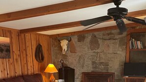 Un salon chaleureux avec une cheminée en pierre, des poutres apparentes, un canapé rouge, une table basse en bois et un ventilateur de plafond.