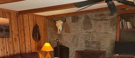 A cozy living room with a stone fireplace, wooden beams, a red sofa, a wooden coffee table, and a ceiling fan.