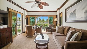 A spacious living room with a ceiling fan, a large window with a view of palm trees, a brown sofa, and a coffee table. - F303 - 2 Bedroom Garden Value Suite in Kapaa - Steps from the Water! AC Comfort! (Kapaa)