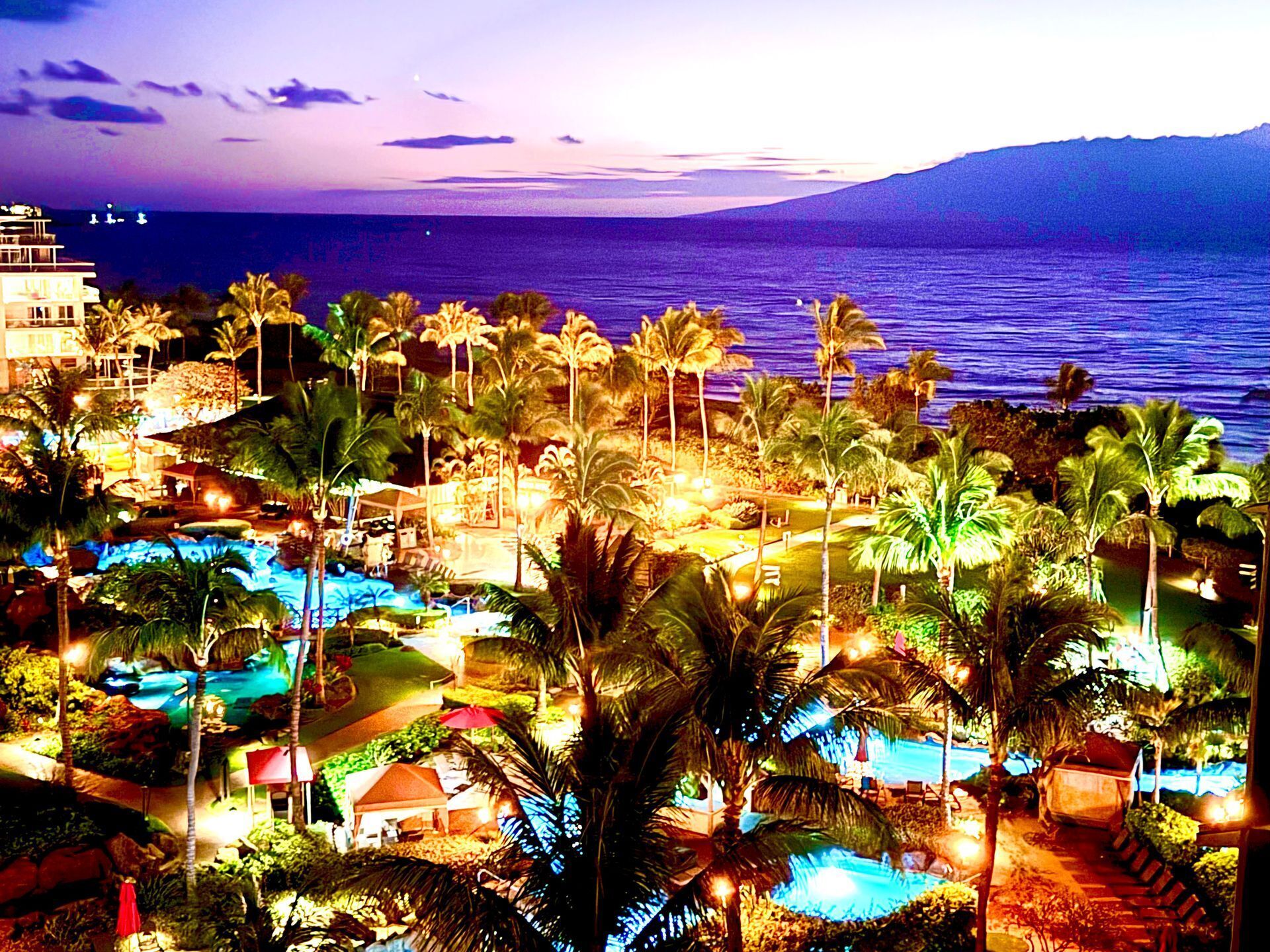 A resort with multiple swimming pools, palm trees, and a view of the ocean at dusk.