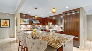 A modern kitchen with a marble countertop, stainless steel appliances, and a central island with seating. - Maui Resort Rentals: Honua Kai Konea 749, Premier 7th Floor Penthouse 3BR w/ Gorgeous Ocean and Black Rock Views! (Lahaina)