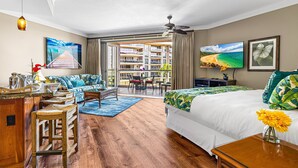 Ein Hotelzimmer mit einem großen Bett, einem Schreibtisch mit Stuhl, einer Couch, einem Balkon mit Blick auf das Hotel und den Strand sowie einem Deckenventilator.