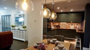 A modern kitchen with a dining area. It includes a wooden dining table set with plates, bowls, and cutlery. There are hanging pendant lights above the table. The kitchen features a brick backsplash and stainless steel appliances. - NEW Modern 4-Bed House, Deluxe Living, 3 Bathrooms (Reading)