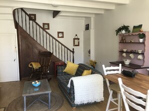Una sala de estar acogedora con una escalera de madera, un sofá a cuadros con cojines amarillos, una mesita con un bol de fruta y una mesa de comedor de madera con sillas.