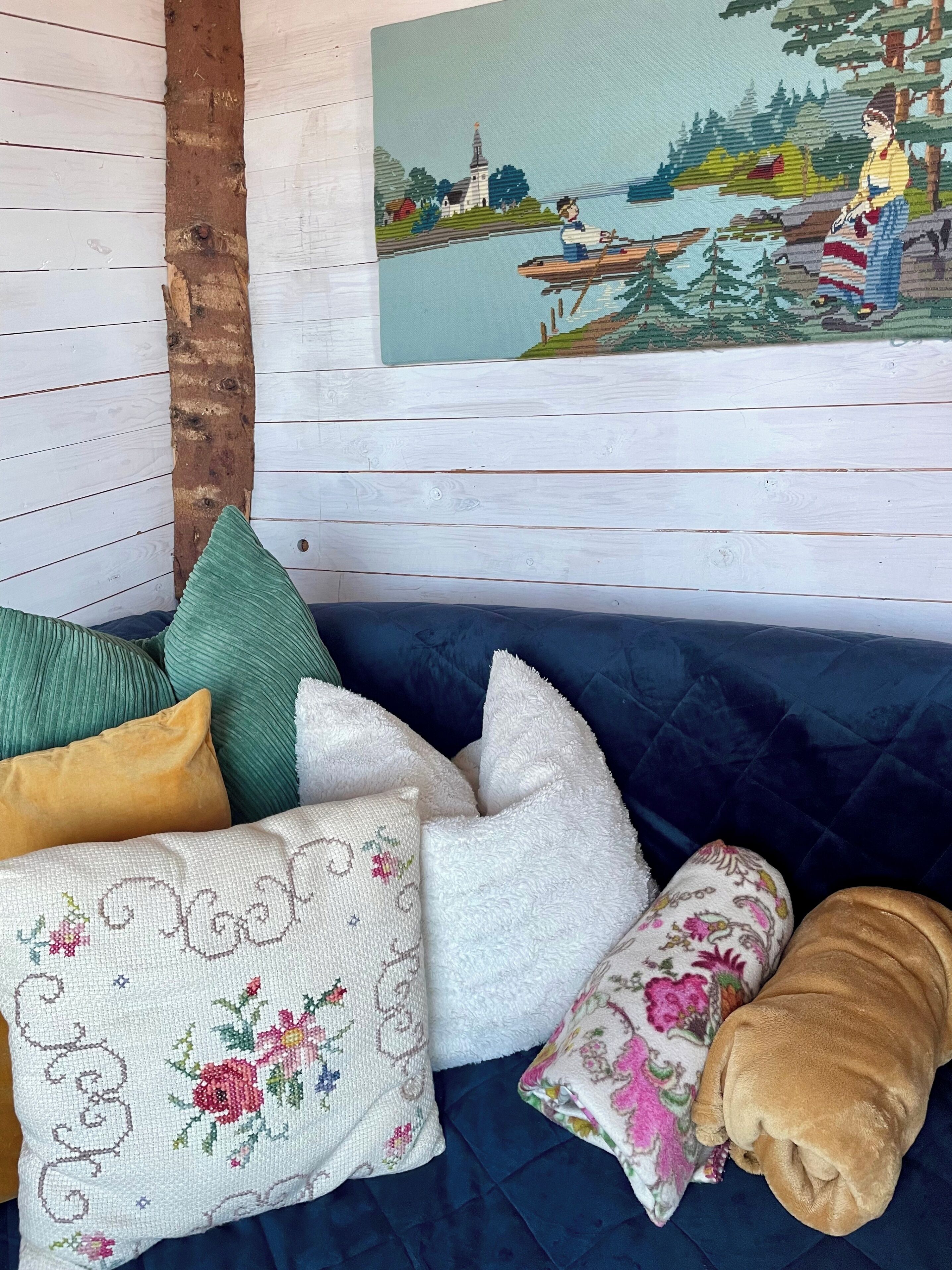 A room with a blue sofa, various pillows, a wooden wall, and a painting of a landscape.