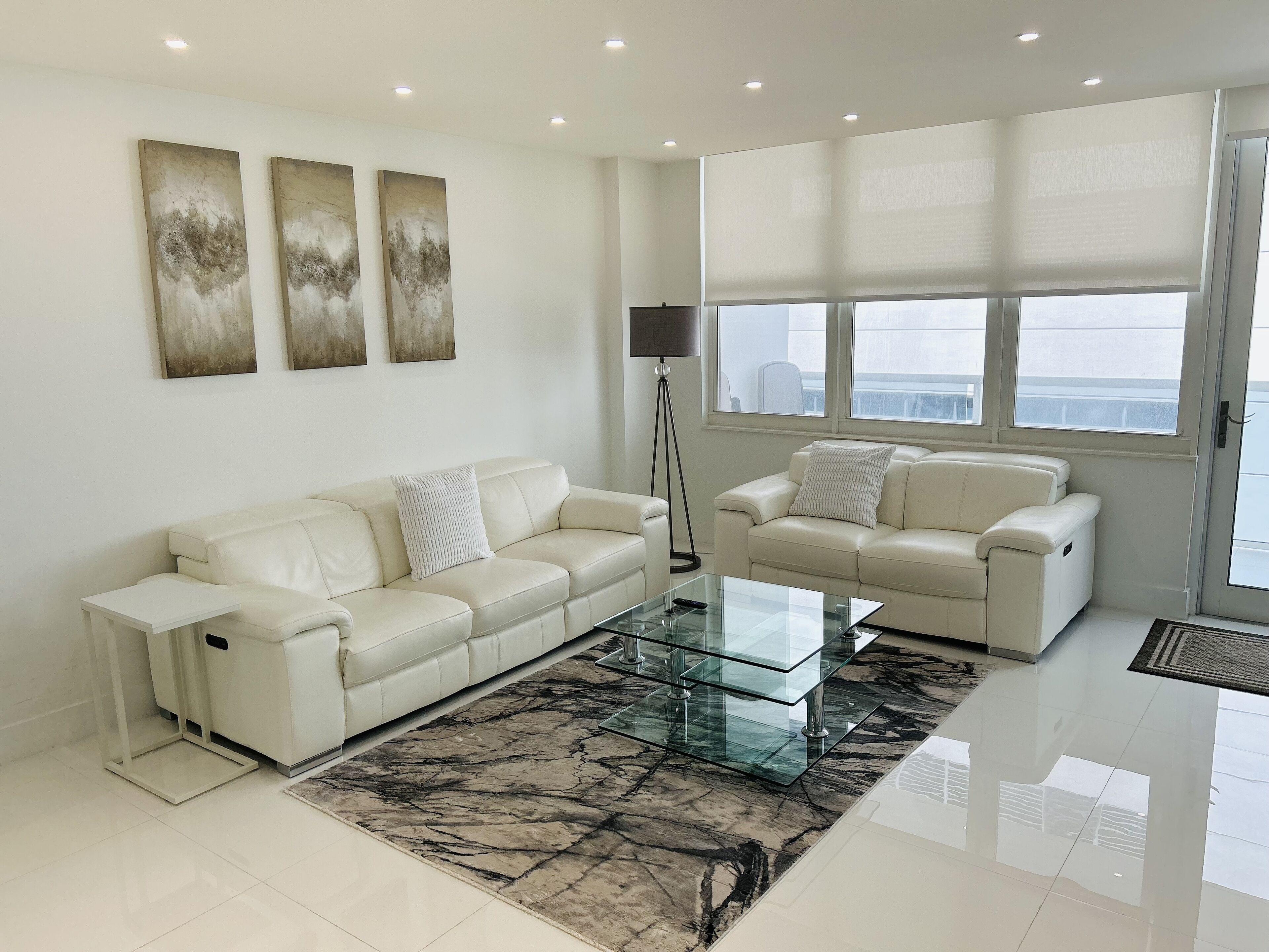A modern living room with two white leather sofas, a glass coffee table, and a large window with blinds.