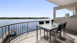 Ein Balkon mit Glastisch und Stühlen, der Blick auf ruhiges Wasser und eine entfernte Küstenlinie bietet.