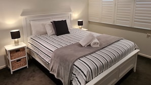 Ein ordentlich bezogenes Bett mit gestreifter Bettwäsche, zwei Nachttischlampen, eine wandmontierte Klimaanlage und ein Fenster mit Jalousien.