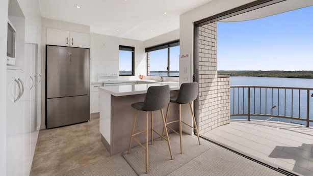 Eine moderne Küche mit einer großen Kochinsel, zwei Barhocker und Blick auf einen See durch einen Balkon.