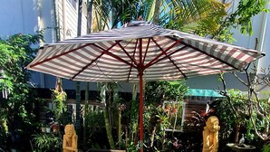 An outdoor patio with a striped umbrella, wicker furniture, and a decorative statue. - Cotton Tree Tranquil Gem (Maroochydore)