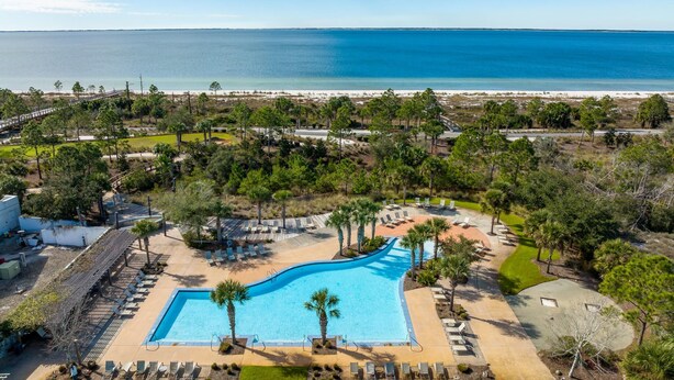 Luchtbeeld van een resort met een centraal zwembad, omgeven door palmbomen en loungegedeeltes, met een strand en oceaan op de achtergrond.