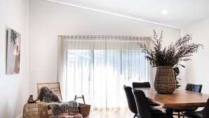 Un coin repas comprenant une table en bois, des chaises noires, une petite table d’appoint en osier et un grand vase tressé contenant des plantes.