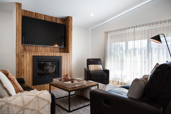 A modern living room with a flat-screen TV mounted on a wooden wall, a fireplace, a comfortable sofa, and a large window with sheer curtains.