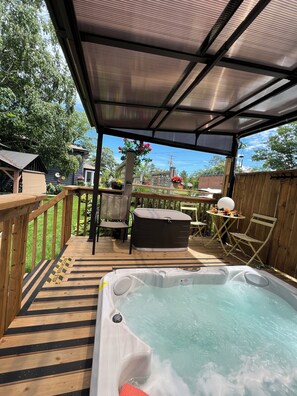 Una terraza de madera con un jacuzzi, muebles de exterior y un espacio cubierto con vistas al jardín trasero.