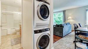 Ein Waschraum mit gestapelten Waschmaschinen und Trocknern, ein Badezimmer mit Toilette und Dusche sowie ein Wohnbereich mit Sofa und Tisch.